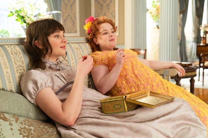 Bridgerton Eloise and Penelope sat next to on another on a couch, eating chocolate