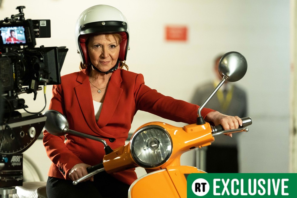 Doctor Who Bonnie Langford exclusive Bonnie Langford sits atop a yellow Vespa with a white helmet on and red blazer in a shot from filming Doctor Who in Doctor Who Unleashed. The RT Exclusive sticker sits in the bottom right corner of the image.