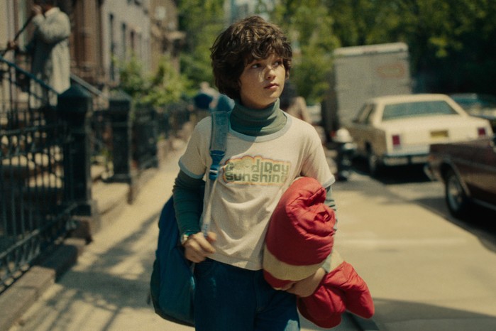 Eric: Limited Series Ivan Howe as Edgar in Eric wearing a Good Day Sunshine t-shirt and walking down a street holding a red jacket and blue bag.