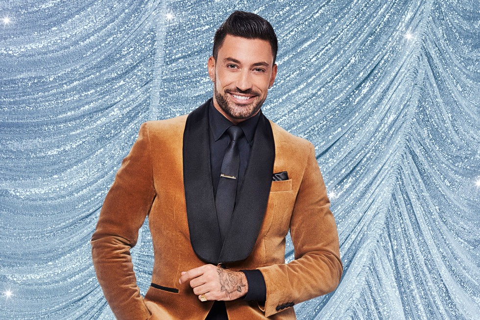 Giovanni Pernice Giovanni Pernice smiling in front of a blue curtain background. He is wearing a brown velvet suit with one hand in front of him and the other in his pocket.