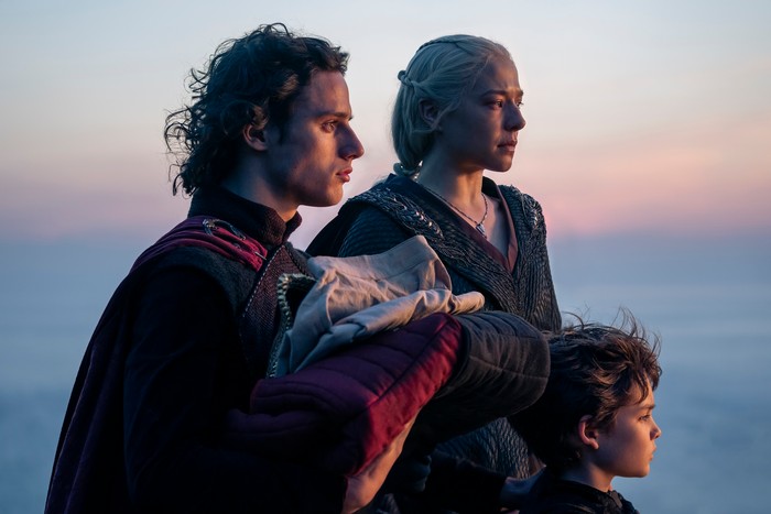 harry-collett-emma-d-arcy-oscar-eskinazi Harry Collett as Prince Jacaerys Velaryon holding a baby, Emma D'Arcy as Queen Rhaenyra Targaryen and Oscar Eskinkanzi as Prince Joffrey Velaryon stare out to sea in House of the Dragon season 2.