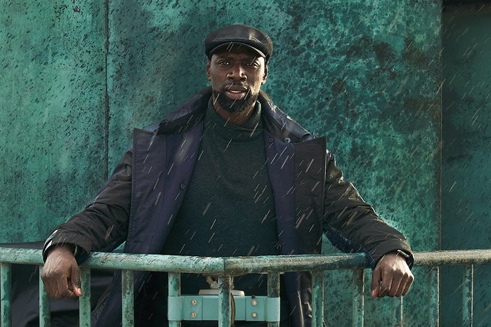 Lupin Season 3 Assane Diop in Lupin wearing a dark jacket and cap, leaning on a rail