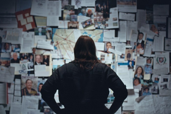 The_Program_Cons_Cults_Kidnapping_Netflix Katherine Kubler standing with her back to the camera and hands on her hips standing in front of a board filled with evidence and photos in The Program: Cons, Cults and Kidnapping.