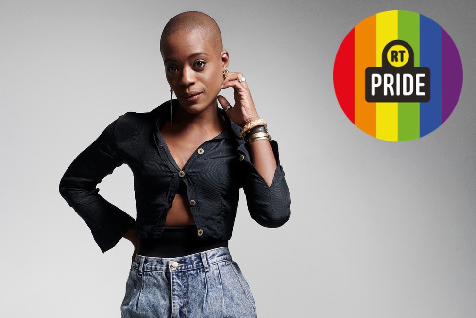 T'Nia Miller T'Nia Miller looking at the camera standing against a white background. She's wearing jeans and a black top. In the upper right corner, a rainbow badge reads: RT Pride.